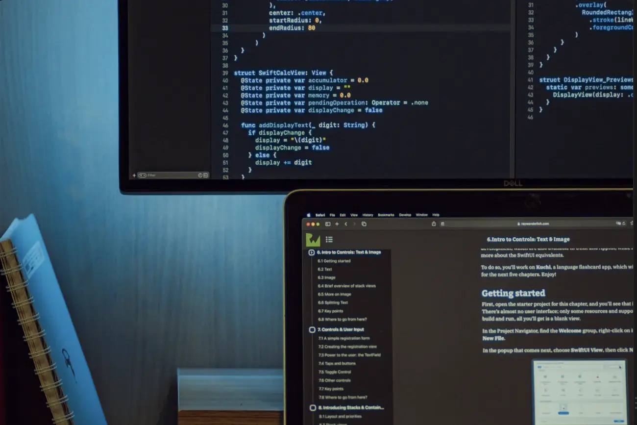 A desk lit blue at night — a monitor of Swift source standing behind a laptop open on developer documentation, notebooks stacked at the left.