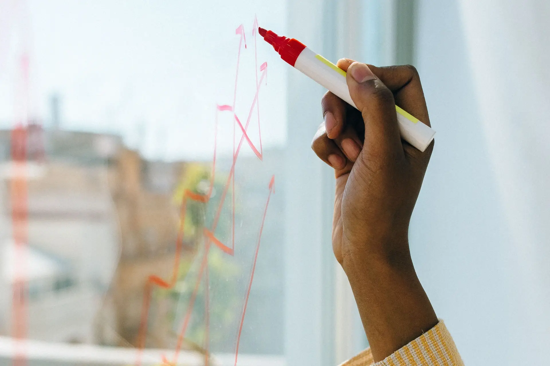 A hand drawing a rising line in red marker across a glass panel, over the street outside.