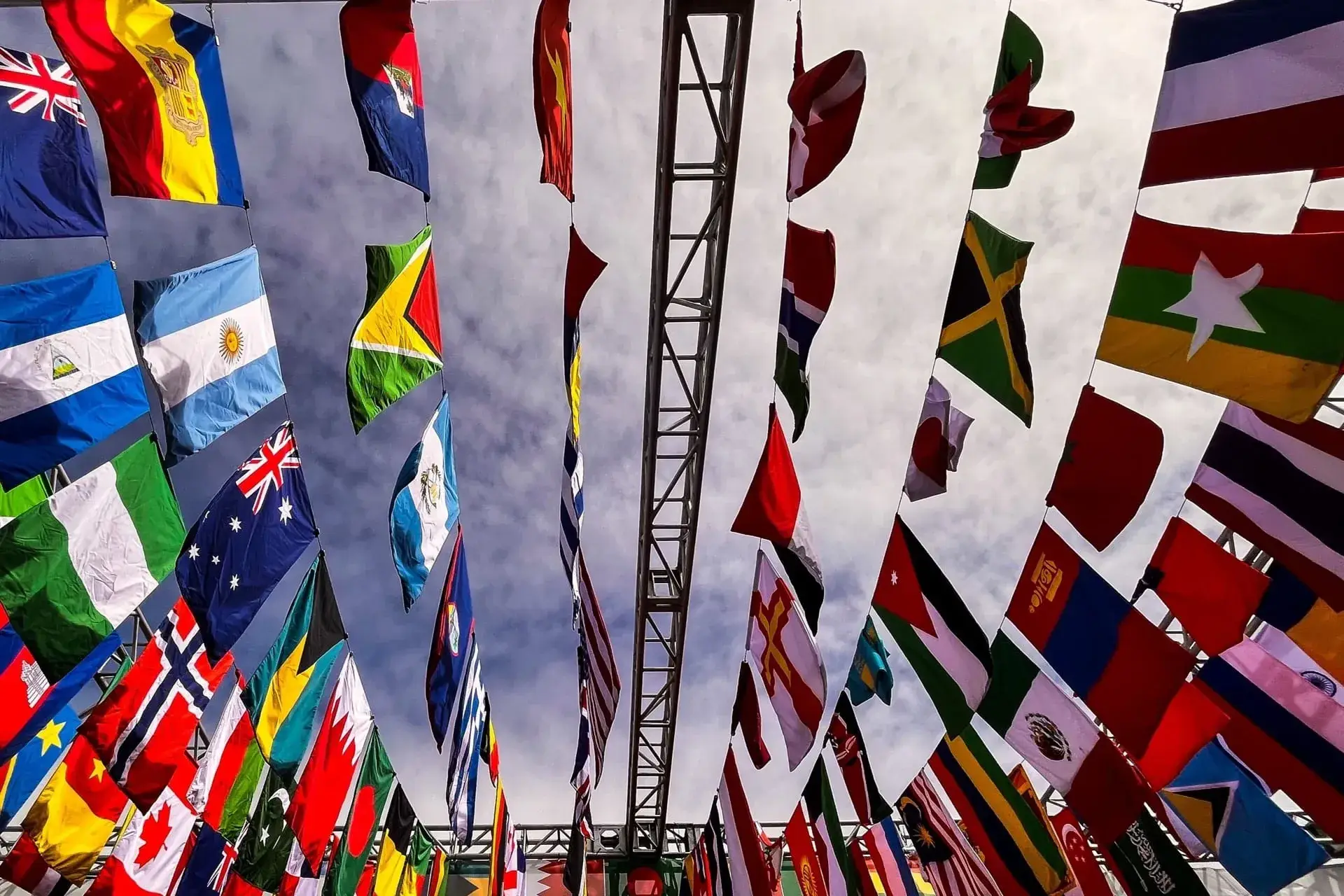 Rows of national flags hanging from a frame against an overcast sky, seen from below.