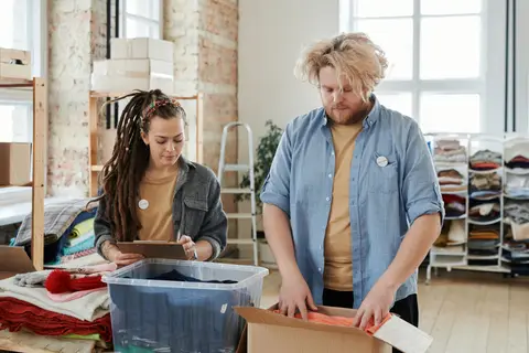 Two people packing orders in a small studio, stock shelved on the wall behind them.