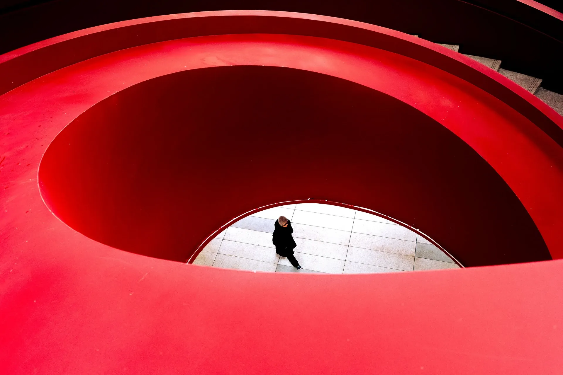 Looking down through the openings of a red spiral structure at a single person crossing the floor below.