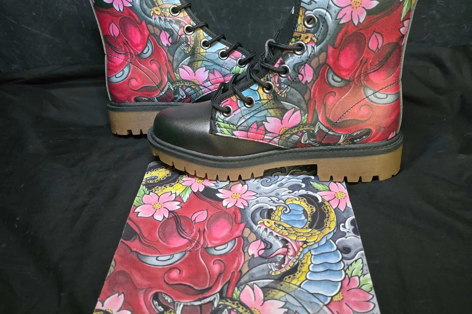 A pair of TattooMyShoes boots printed with a red hannya mask and snake design, photographed on black beside the artist's original hand-painted flash panel it was taken from.