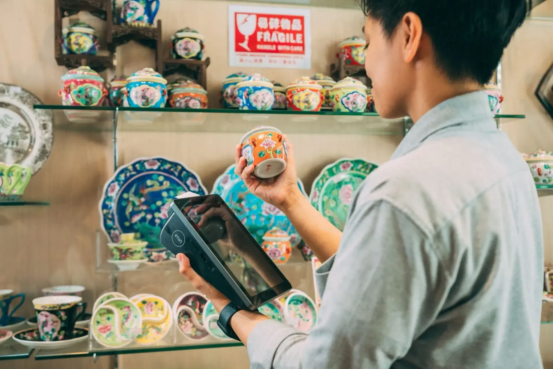 A shop assistant holding a tablet, checking one piece of stock against a shelf of decorated ceramics.