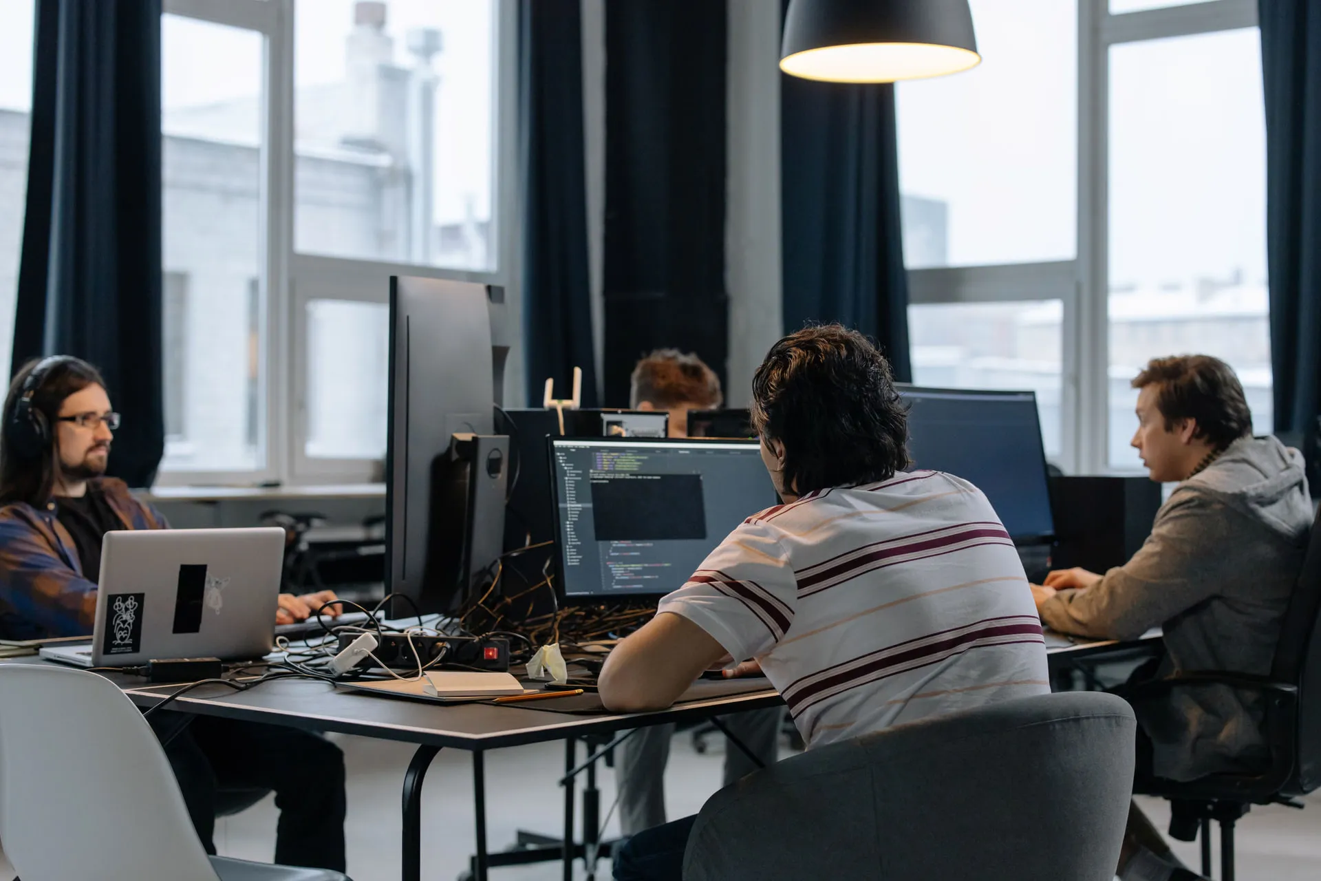 Three developers at a shared studio desk in front of tall windows, one with code open on a monitor