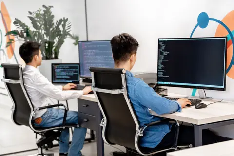 Two developers working side by side at adjacent desks, theme code on the monitors in front of them.