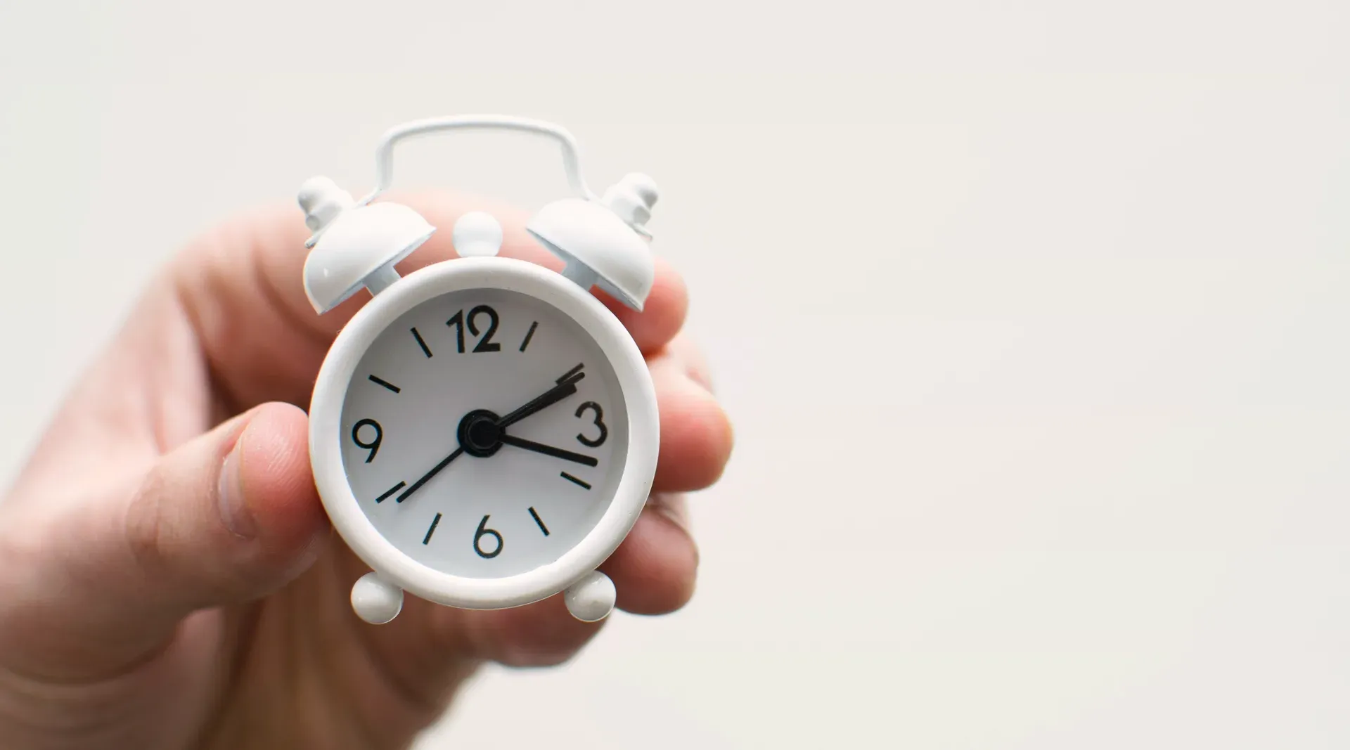 A hand holding out a small white alarm clock against a plain pale ground.
