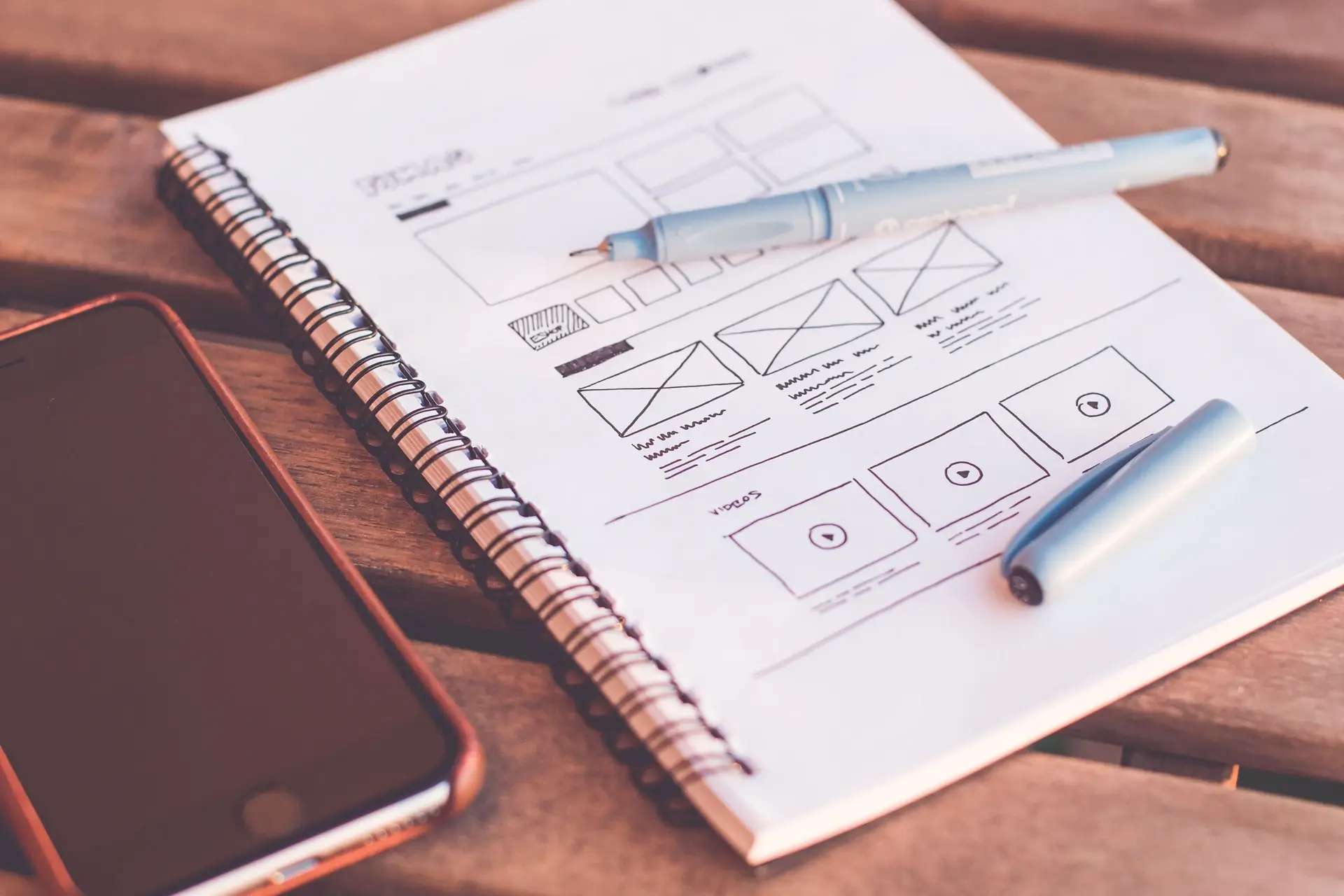 A spiral sketchbook open on a desk, its page drawn with wireframe blocks for a header, a grid and a row of tiles, a pen resting across it.